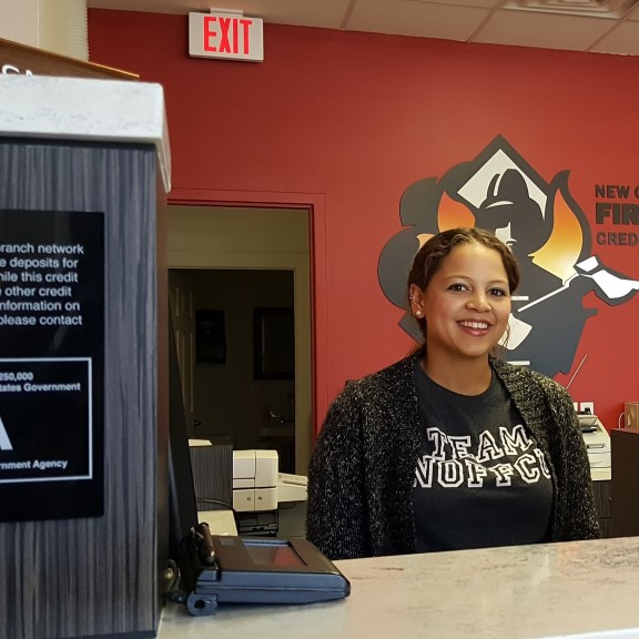 A teller at NOFFCU stands smiling behind the counter wearing a TEAM NOFFCU shirt