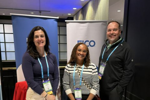 3 people who work at FICO stand behind an exhibit table, smiling