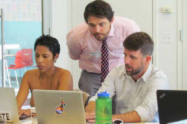 An instructor consults with two CU staff members working on laptops